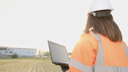 A worker stands in a field looking at a laptop. The sun is rising in the background. A factory can be seen in the distance. The worker is wearing safety gear © Happy Photo