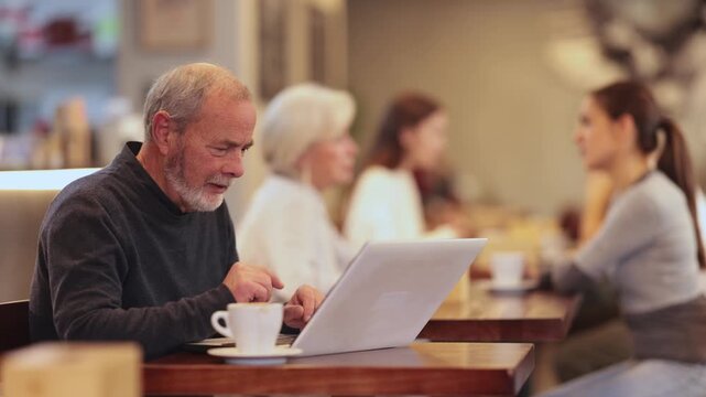 During lunch break, senior woman spends time with laptop at table in cafe, drinks coffee, and communicates via video link with relatives. Freelance senior woman using laptop and drinks coffee. 