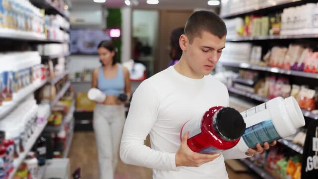  Guy choose protein sports nutrition in store, enriched protein shake, carefully examines label on package. Shop visitor reads composition and characteristics of protein shake