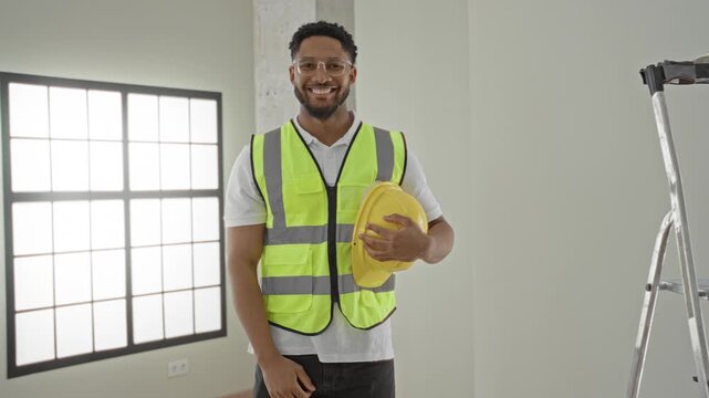 Man architect wearing high visibility vest holding yellow hardhat to chest and smiling inside building with ladder and window; professional pride.