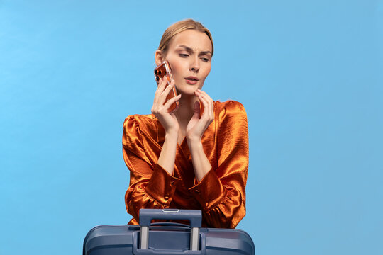 Blonde woman in orange blouse holding phone near face looks concerned, standing behind suitcase on blue background. Concept of travel, communication, and uncertainty in journey planning.