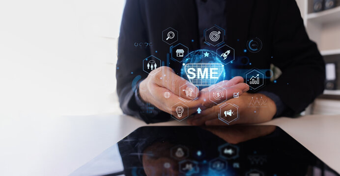 Person in a dark suit holding hands presenting a glowing sme globe surrounded by a network of hexagonal business, marketing, and growth icons, with a tablet reflecting the visuals on a bright desk
