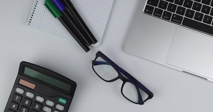 Top down flat lay of a silver laptop and calculator with pencils plus notebook as a hand adds blue glasses on white background now<