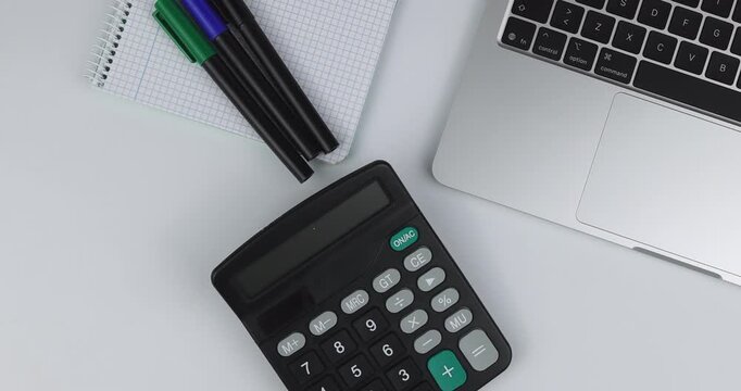Overhead view of a silver laptop with pencils and notebook on a white background as a hand places a black business calculator here.