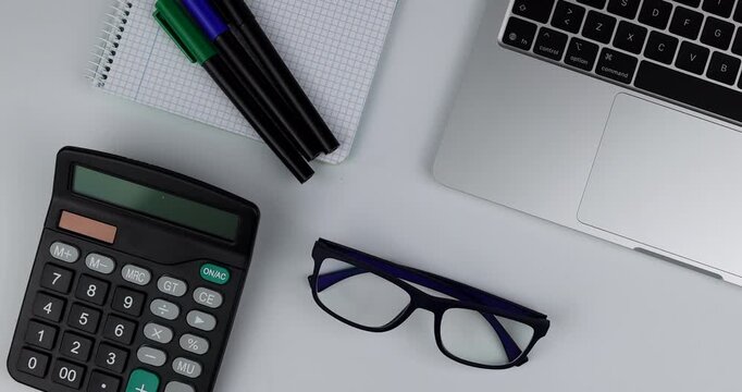 Overhead view of a silver laptop with calculator plus pencils and notebook as a hand places blue glasses on a white background set