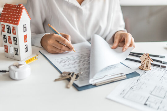 Person reviewing real estate documents with blueprint, keys, miniature house on table. Signing of mortgage contract. Buying apartment, signing agreement, renovation planning and secure home investment