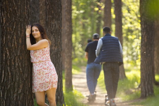 A young woman walking in the forest. A beautiful woman resting in a forest, leaning against a pine tree trunk. Behind her, a group of guys on scooters rides along a path.