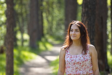 Fototapeta premium Portrait of a young woman in a park. A woman stands on a path between pine trees and looks away. The background is beautifully blurred.