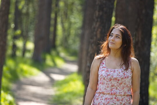 Portrait of a young woman in a park. A woman stands on a path between pine trees and looks away. The background is beautifully blurred.