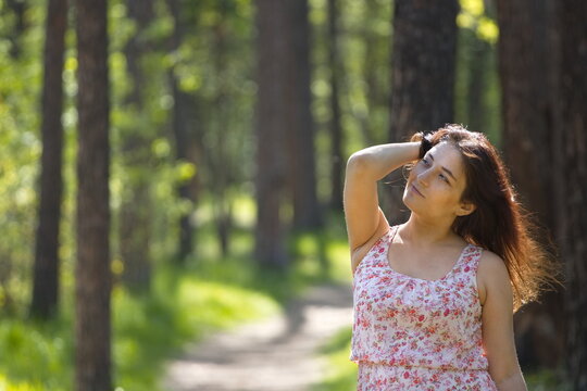 Portrait of a young woman in a park. A woman stands on a path between pine trees, looking away, adjusting her hair with her hand. The background is beautifully blurred.