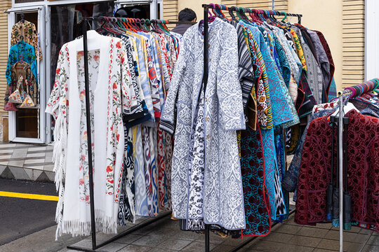 Traditional Uzbek robes and textiles displayed on racks at Siab bazaar in Samarkand. Colorful ikat and embroidered garments for sale at an outdoor market, ideal for travel and cultural content.