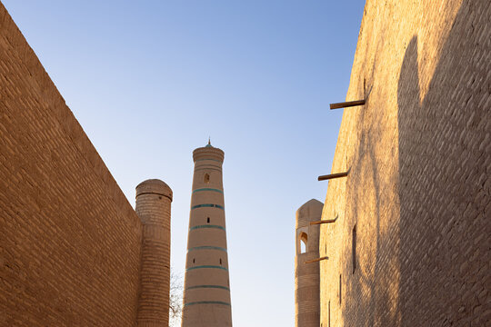 Minaret rising above mud-brick walls in the Ichan Kala old town of Khiva, Uzbekistan. Warm golden light highlights the ancient brickwork against a clear blue sky. Ideal for travel publications.