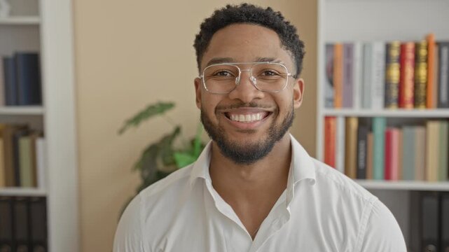 Man wearing glasses smiling directly at camera in a study building with bookshelves and plant visible; confidence success.