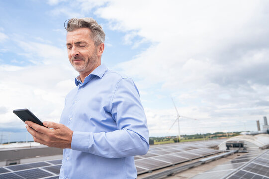 Smiling businessman using smart phone on rooftop under cloudy sky