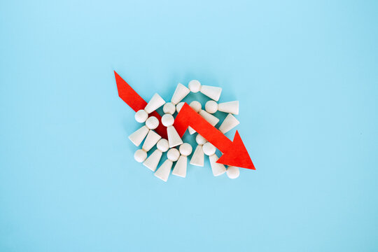 Red downward arrow lies on pile of wooden peg dolls over blue background. Mass layoffs 2026, workforce reduction impact, global unemployment surge, Economic recession.