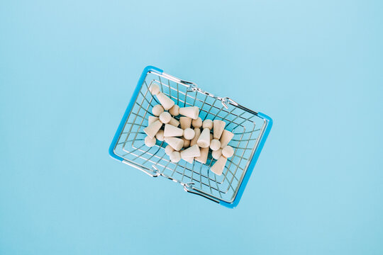 Small wooden peg dolls fill miniature wire shopping basket on bright blue surface. Cost of living crisis, inflation impact on families, basic needs affordability, Consumer purchasing power.
