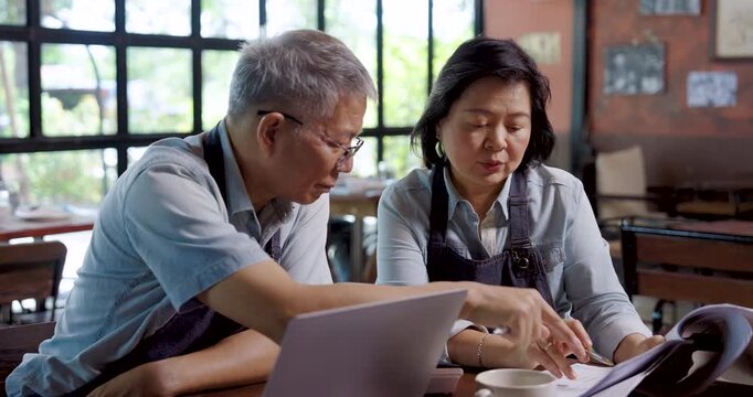Asian senior couple cafe owner wearing aprons sitting at wooden table analyzing financial documents with laptop, teamwork management showing cooperation discussion problem solving in family restaurant