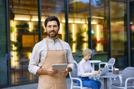 A smiling man wearing an apron holds a tablet while standing outside a restaurant with a blurred background of a patron working on a laptop