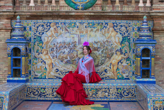 A beautiful flamenco dancer sitting on a tiled bench in a square in Seville, Spain. She is wearing a red ruffled dress and has a flower in her hair. Flamenco, a UNESCO Intangible Cultural Heritage.