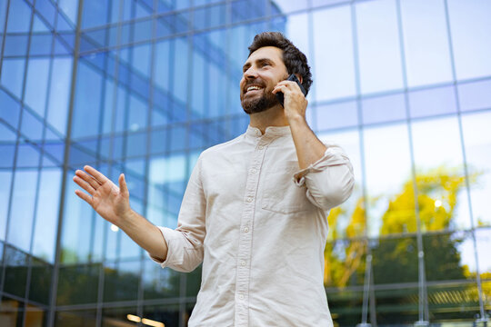 A smiling man with a beard stands outside a modern glass building while talking on his smartphone and gesturing with his hand