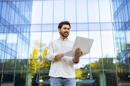A smiling man with a beard stands outside a modern glass building holding a laptop and looking at the screen
