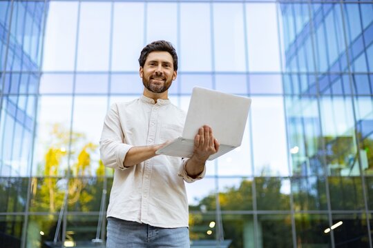 A smiling man holding a laptop stands in front of a modern glass office building with trees reflected in the windows