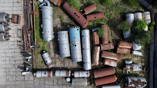 Aerial top-down view of an industrial scrap yard filled with various steel storage tanks, pressure vessels, and cylindrical containers in different stages of decay, rust, and oxidation on concrete gro