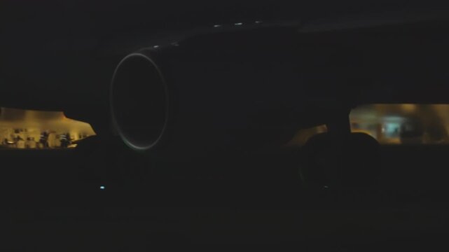 Jet engine nacelle seen from directly below at night, circular intake centered in frame, warm yellow airport lighting visible on both sides, dark atmospheric mood