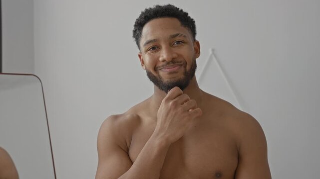 Young man shirtless with bare chest, hand on chin touching beard while smiling in studio next to mirror and wall hook; confidence wellbeing.