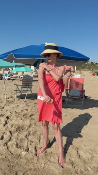 Senior woman aged 80 applying sunscreen on the beach during summer vacation, standing on warm sand in sunhat and coral wrap dress, vertical skincare lifestyle video