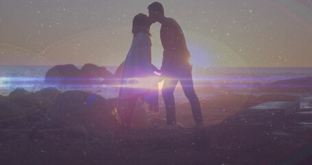 Naklejka premium Holding hands, couple in flowing dress and trousers touching on rocky beach at sunset, lens flare