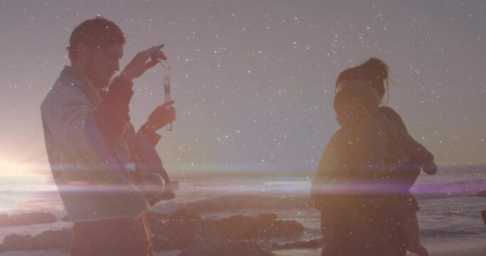 Checking father holding phone while mother holding baby, wearing coats on shore at dusk, lens flare