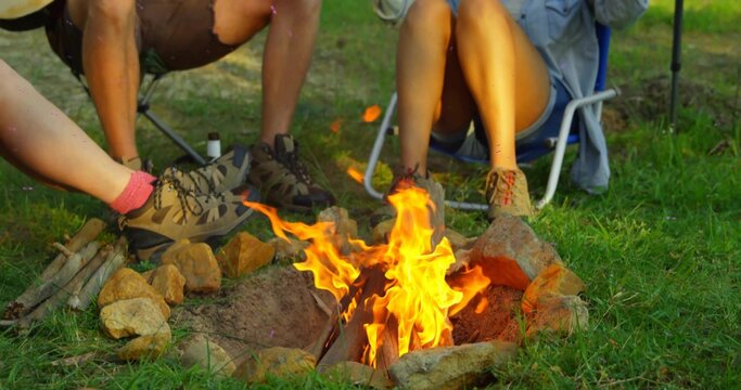 Sitting three adults warming feet by campfire on grassy lawn, with folding chairs and hiking shoes