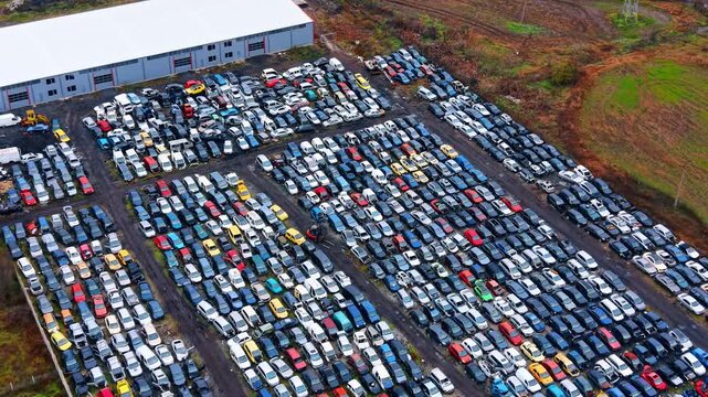 Diagonal aerial perspective of car scrap yard and warehouse. Rows of wrecked cars parked in a salvage facility near a large gray industrial building.