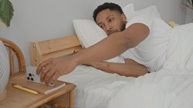 Man reaching with hand for smartphone on bedside table in bedroom with white bedding and light wood headboard; sleepy morning routine.