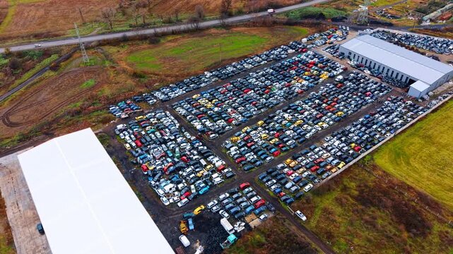 Aerial panoramic view of automotive junkyard and surrounding fields. High angle shot showing a vast collection of scrap cars, an industrial warehouse and country roads.