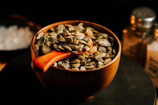 Bowl of raw peeled pumpkin seeds with wooden spoon on a dark surface at home kitchen