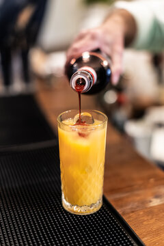 Close-up of red syrup being poured into an orange juice cocktail over ice on a bar mat during event drink service