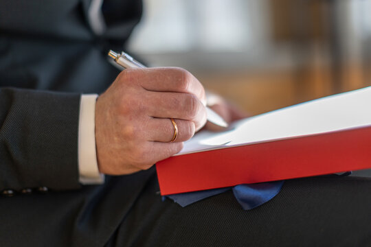 Close-up of a groom wearing a gold ring while signing a document on a red folder during a wedding ceremony