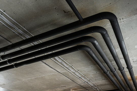 Black insulated pipes run along a concrete ceiling, secured with metal clamps; alongside are white electrical conduits, creating an industrial look.