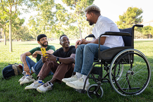 Young African American man in a wheelchair and his male friends laughing and having fun sitting on the grass in a park.