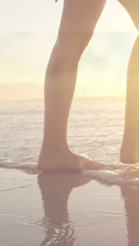 Vertical video: Sun pulling woman to wet sand, barefoot walking, sundress catching light for travel