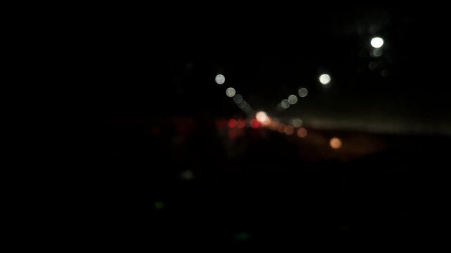 Blurred video background of handheld night drive footage shot from inside a car during rain. Raindrops on the windshield create cinematic bokeh lights from traffic and street lamps, capturing a moody.