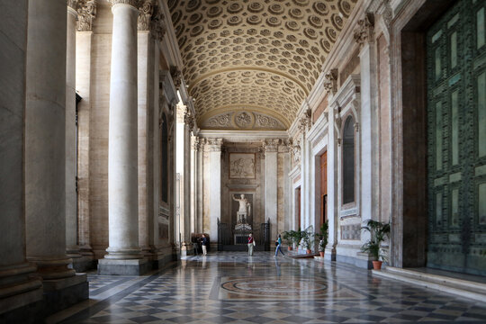 Ampia veduta prospettica del portico monumentale della Basilica di San Giovanni in Laterano con soffitto a cassettoni geometrici, pavimento in marmo riflettente e statua di Costantino, Roma, Italia.