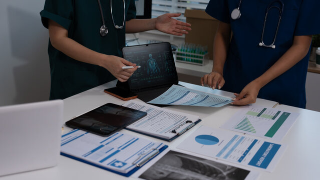 Medical team in scrubs reviewing patient charts and lab reports on desk with laptop showing digital anatomy scan in hospital office.