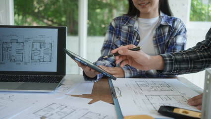 Architects reviewing house floor plans at desk, laptop and tablet in bright office, collaborating on home design project with blueprints and documents. © NanSan
