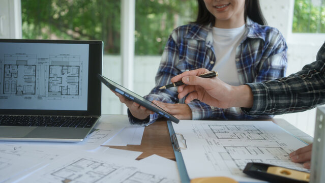 Architects reviewing house floor plans at desk, laptop and tablet in bright office, collaborating on home design project with blueprints and documents.
