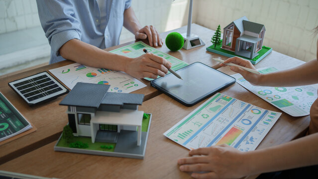 Two colleagues review a digital tablet beside laptops and renewable energy models in a bright modern office, suggesting clean technology planning and teamwork.