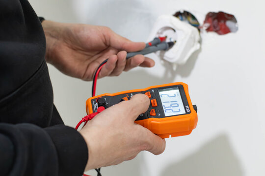 Close-up of a professional technician's hands using a digital multimeter to check the voltage in a power socket during home electrical repair or maintenance. 