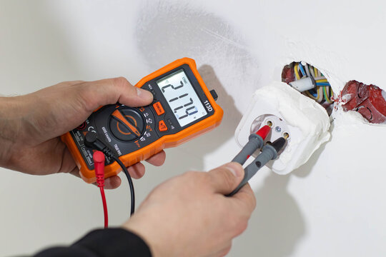 Close-up of a professional technician's hands using a digital multimeter to check the voltage in a power socket during home electrical repair or maintenance. 